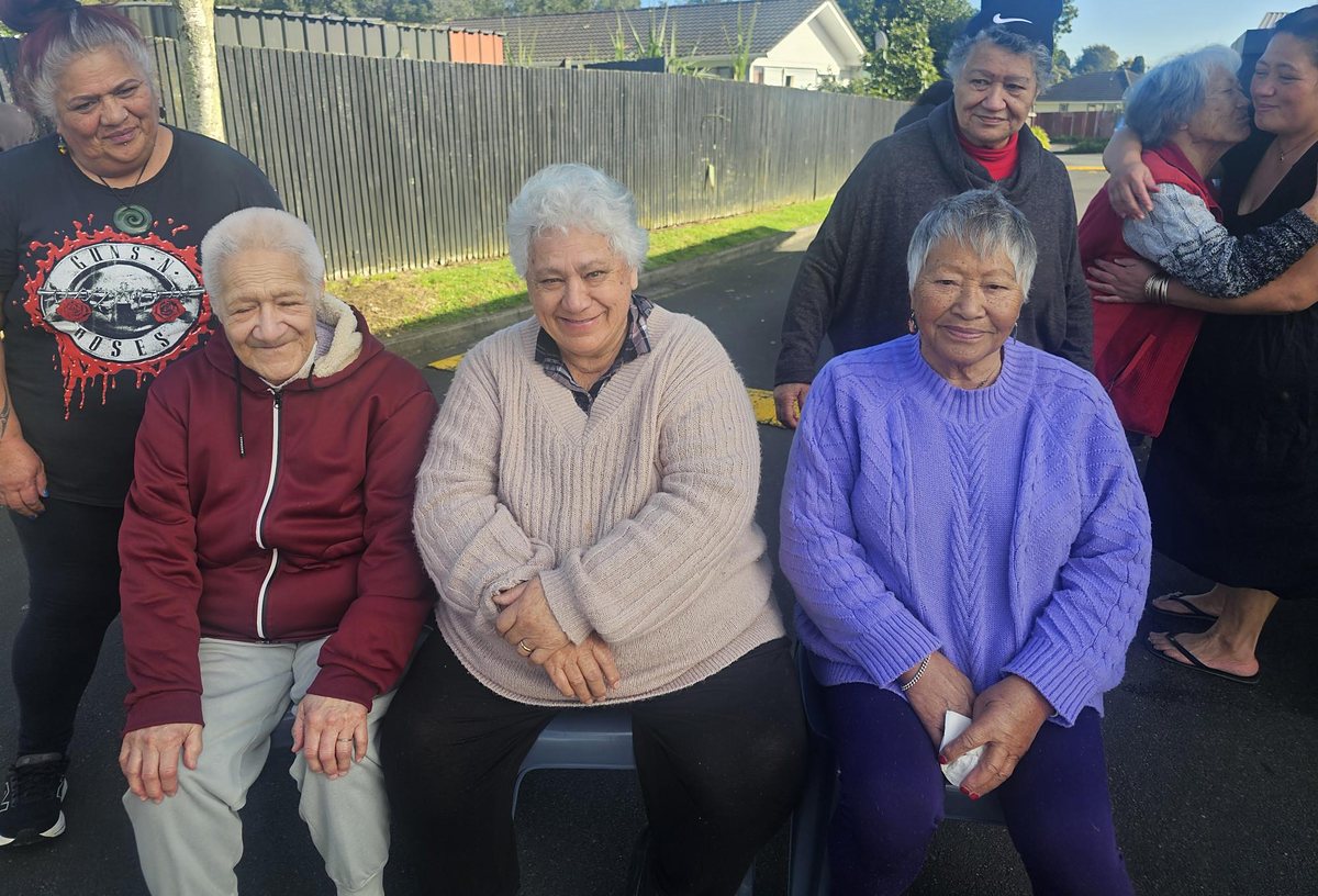 Members sitting together outdoors, smiling, with whānau behind them